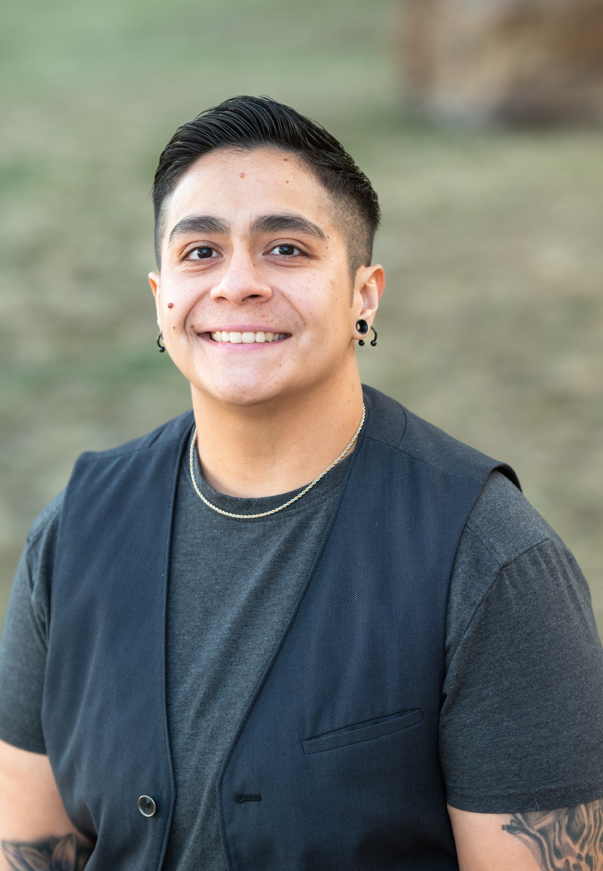 The image features a person smiling at the camera in an outdoor setting with a blurred background of grass. They have short, dark hair styled upwards and are wearing black earrings. The person is dressed in a dark gray tee-shirt layered with a black vest. A thin, gold chain necklace is visible around their neck. The lighting is soft and natural, focused on their face, highlighting an open and friendly expression.  Alt-text:  Person smiling outdoors wearing a dark gray tee and black vest.