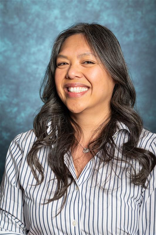 The image depicts a person with long, wavy hair standing against a textured blue background. They are wearing a vertically striped shirt with a blend of white and dark blue stripes. The person is smiling broadly, suggesting a cheerful demeanor. The background features a soft-focus pattern in various shades of blue, providing a subtle contrast to the sharper details of the person in the foreground.  Alt-text:  Person with long hair smiling, wearing a striped shirt against a blue background.