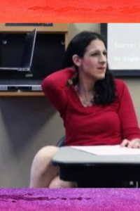 Picture of Dr. Sophia Sangwanthong Director of LGBT+ and Asian & Pacific Islander Program. The image shows a woman with long black hair, wearing a red long-sleeved shirt sitting on a chair at a table in front of a neutral-colored wall. She has one arm resting on the table in front of her and the other arm raised to touch the back of her neck. On the table in front of her is a piece of paper. Behind her, on a shelf attached to the wall, is a closed laptop. To the left of the laptop, some black cables are visible, hanging down. There is a projection screen in the background with faint, unreadable text on it. Surrounding the image is a border with a gradient of bright rainbow colors: red at the top, followed by orange, yellow, green, blue, and purple at the bottom.