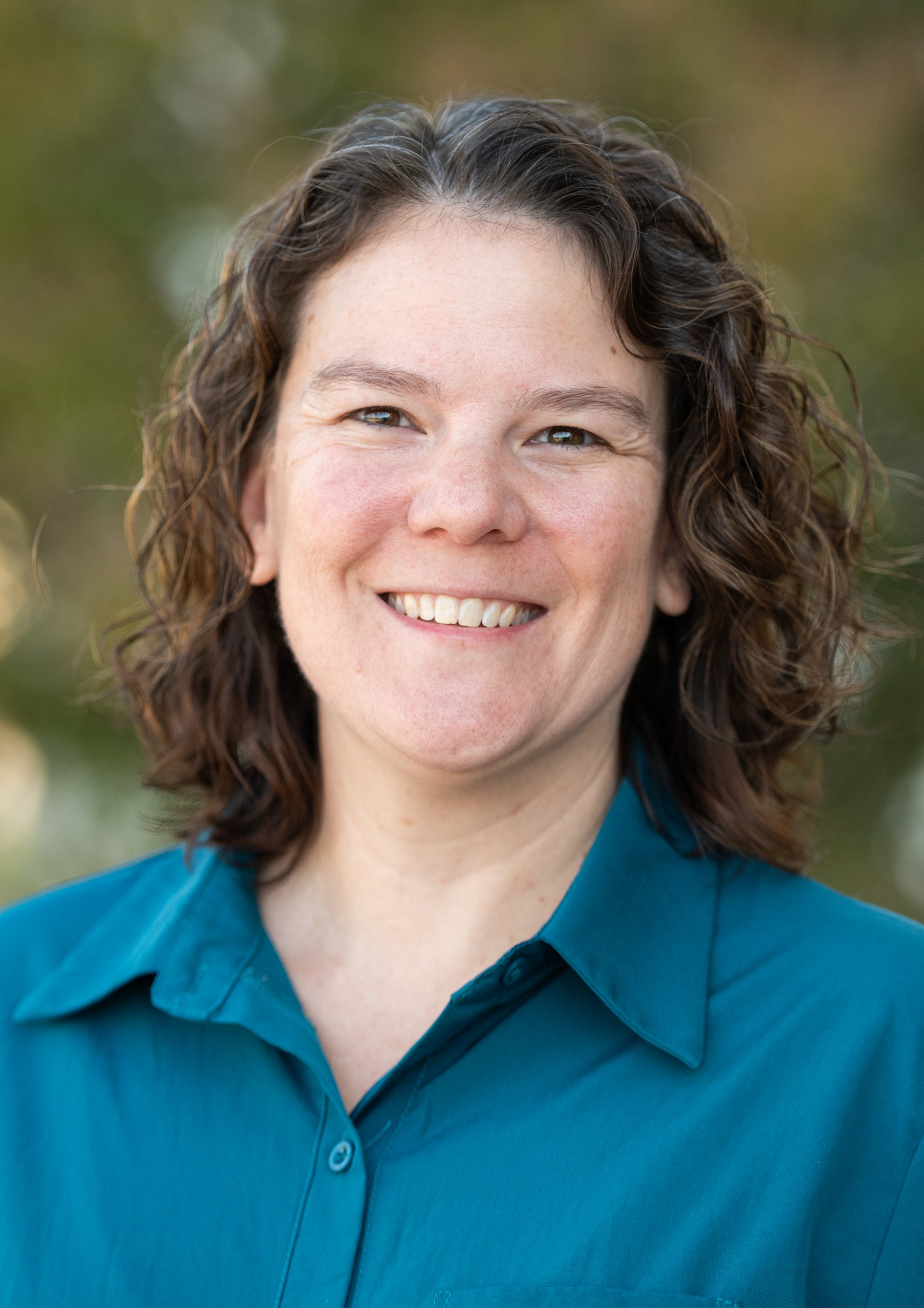 Picture of SPRI AUBREY Administrative Assistant/Special Executive, Office of Equity, Inclusion and Diversity. The image is a head-and-shoulders portrait of an individual against a mottled blue background. The person is facing the camera and smiling slightly. They have straight, short brown hair that is parted to one side. They are wearing a black blazer over a maroon collared shirt. The background has a textured, abstract pattern with various shades of blue and a slightly blurred appearance.