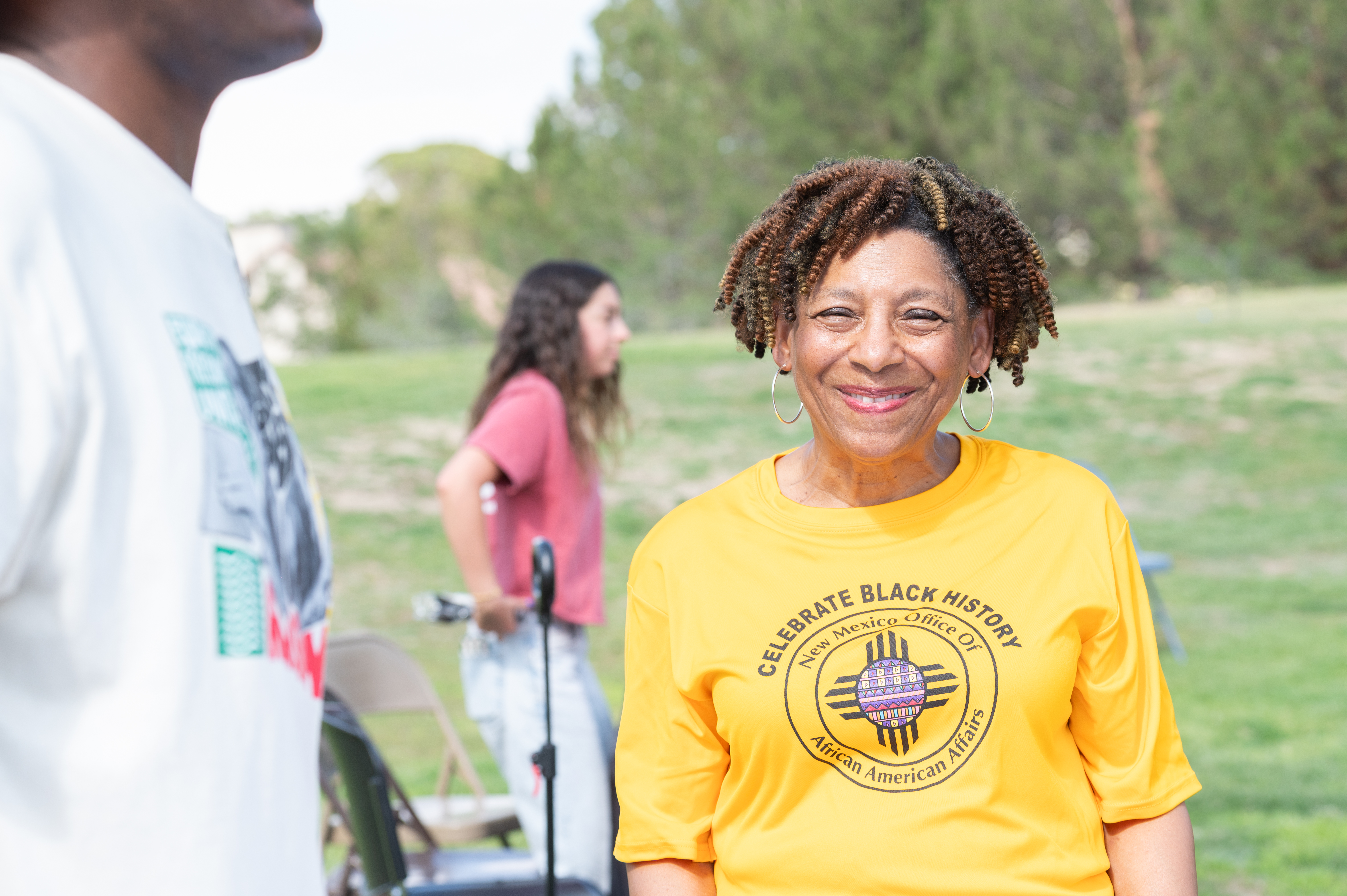 Picture from the celebration of Juneteenth, The image shows an outdoor setting with three individuals present. The primary focus is on a woman in the foreground who is smiling at the camera. She is wearing a bright yellow T-shirt with text and a circular logo on the front. Her hair is styled in short, curly twists, and she is wearing hoop earrings. Behind her to the right, there is another person with long hair, standing sideways and wearing a red T-shirt and light blue jeans. To the left, partially in the frame, is a person with dark skin wearing a white T-shirt. The background features a grassy field and trees.