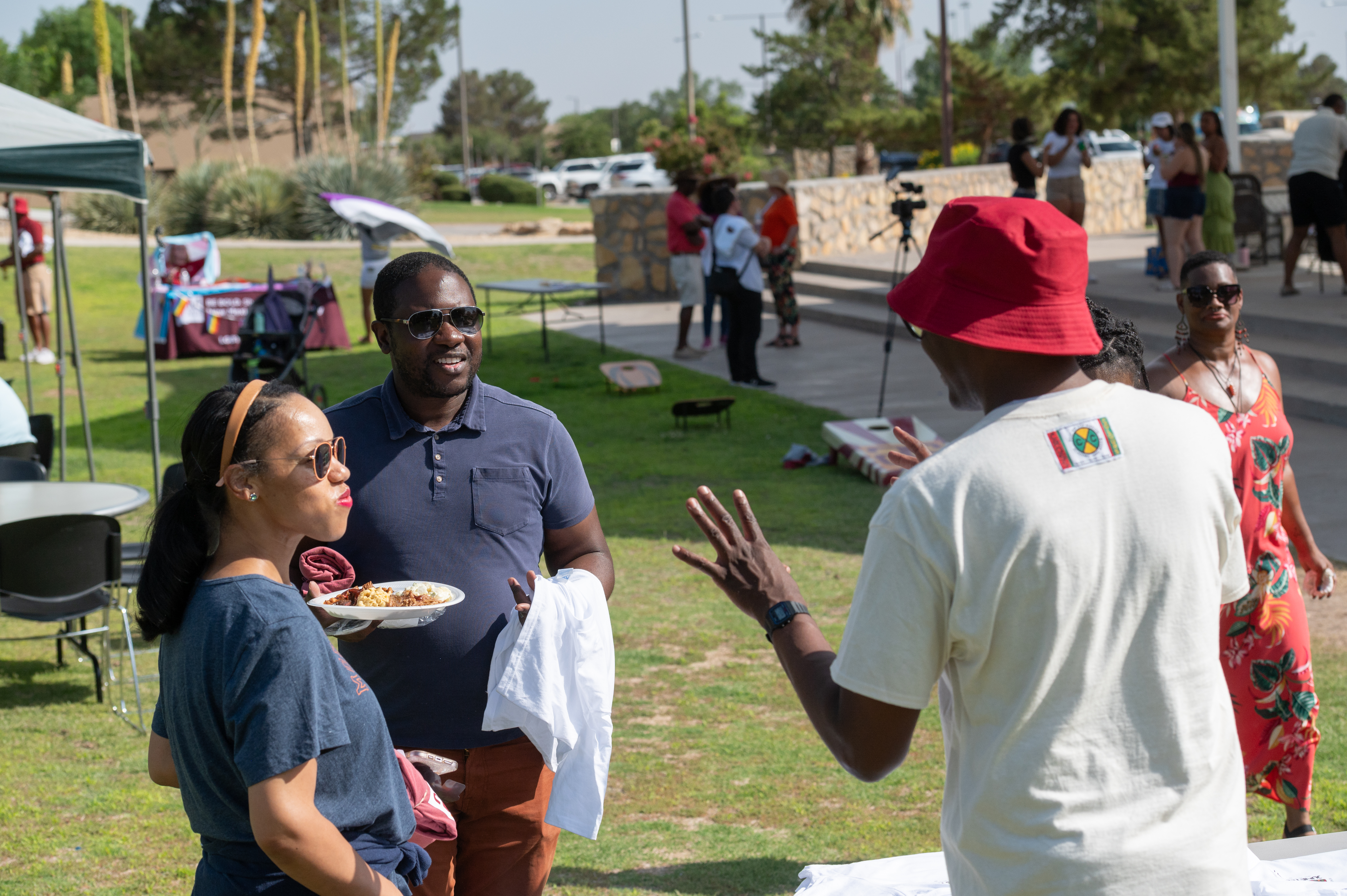 Picture from the celebration of Juneteenth, The image portrays a casual outdoor gathering on a sunny day, possibly a picnic or community event in a park. In the foreground, three people are interacting. A woman with a headband and sunglasses, holding a plate of food, stands facing two men. The man in a navy blue polo shirt and sunglasses appears to be engaged in conversation with another man wearing a white T-shirt with a small multicolored logo on the back and a red bucket hat, who is gesturing with one hand. In the background, other people are scattered around the park, with some standing and others seated. A green canopy tent is set up to the left, alongside tables and chairs. In the mid-ground to the left, another table appears to display an informational banner with unclear text. The park is lush with green grass and has various trees and shrubbery around, with a stone wall and sidewalk in the distance. The sky above is bright and clear.