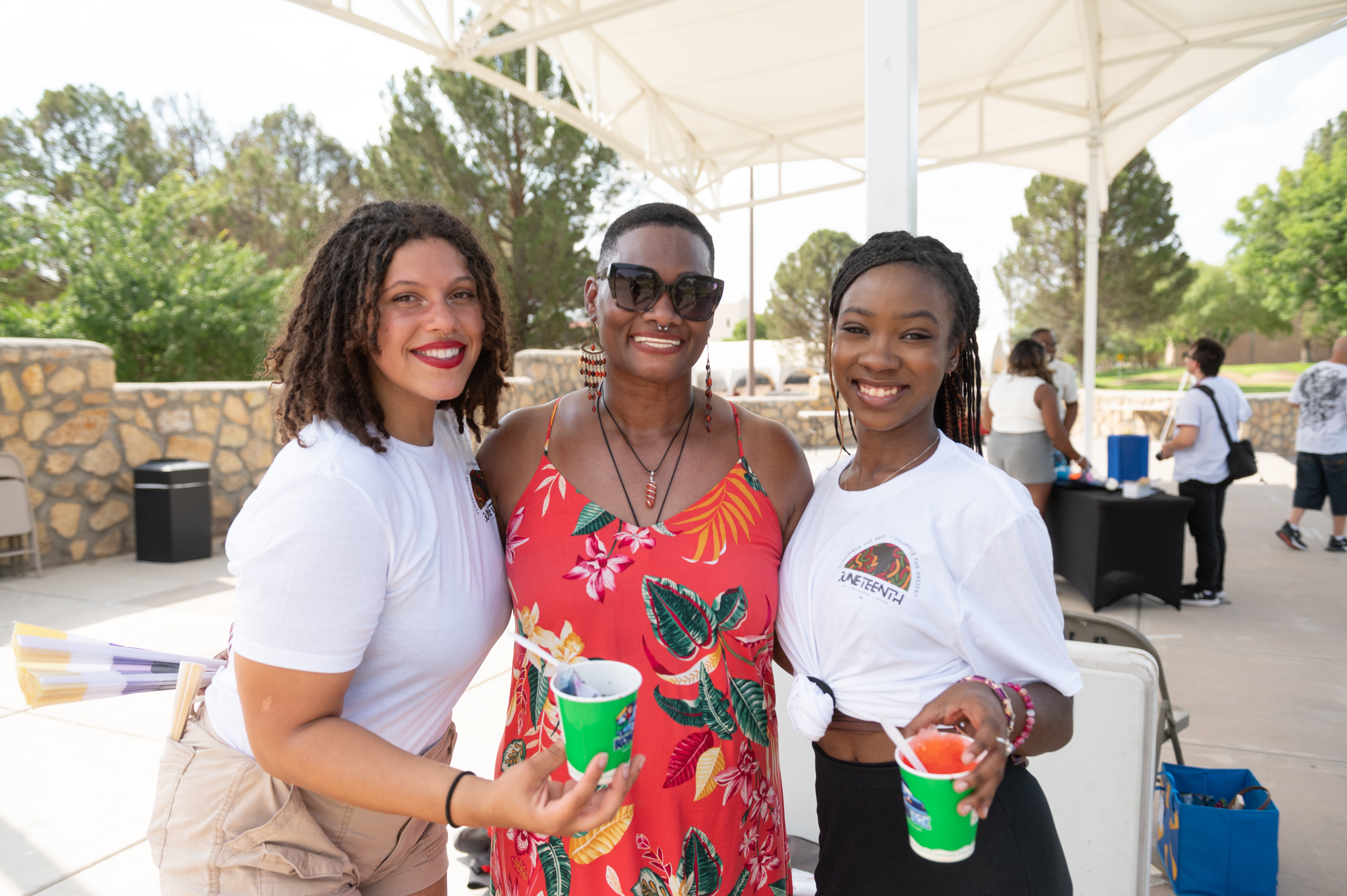 Picture from the celebration of Juneteenth, The image portrays three women standing closely together and smiling at the camera under a white canopy. The woman on the left has curly hair, is wearing a white T-shirt, khaki shorts, and holds a green cup with shaved ice. The woman in the middle has short hair, wears dark sunglasses, a red sleeveless dress with a colorful leaf pattern, and is also holding a green cup with shaved ice. The woman on the right has long braided hair, is wearing a white T-shirt with a visible logo and the word "JUNETEENTH," and is holding a green cup with a colorful drink. Behind them, there is an outdoor area with trees, a stone wall, and several people in the background.