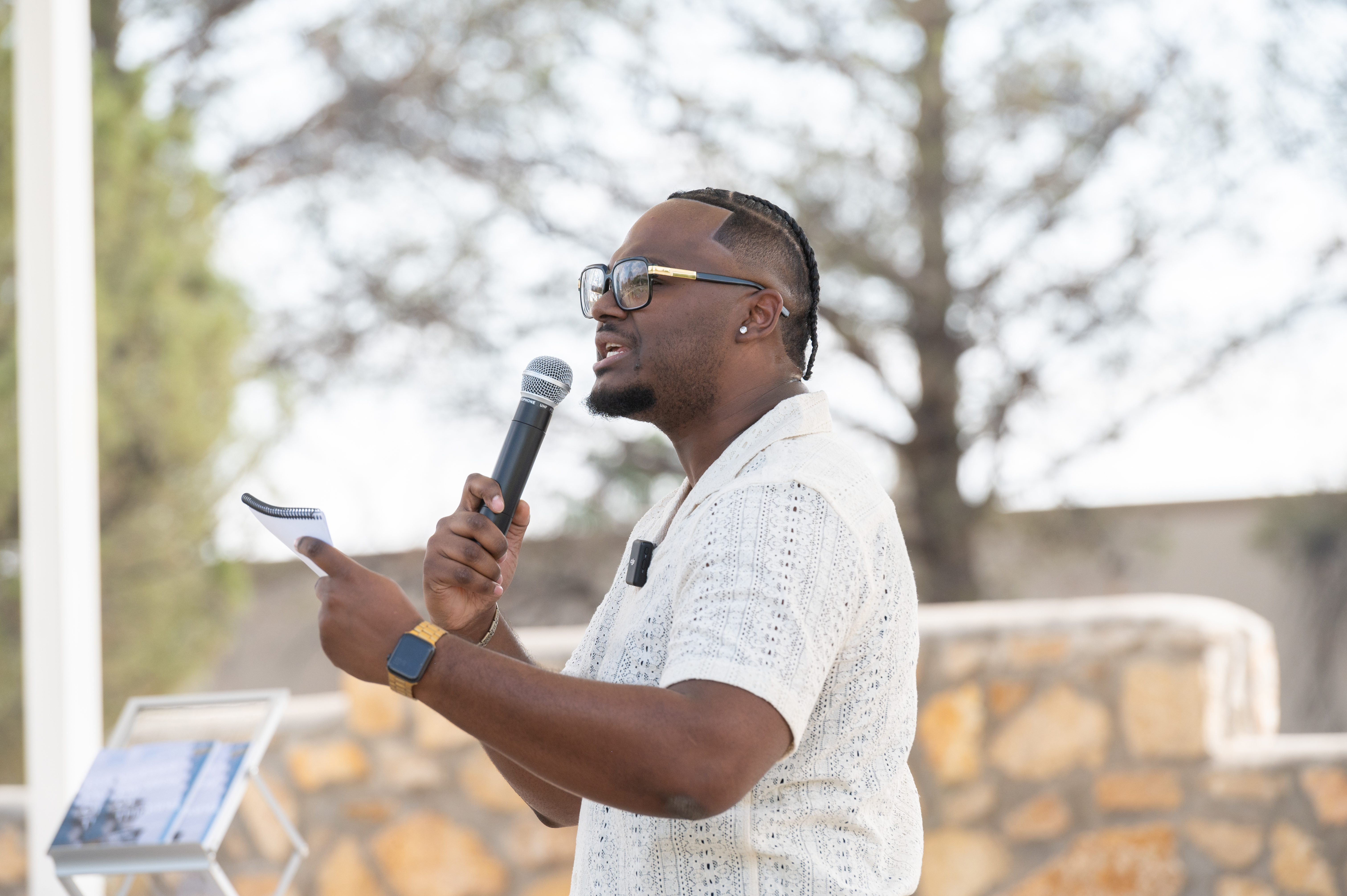 Picture from the celebration of Juneteenth, The image portrays a man standing outdoors, speaking into a microphone with his left hand while holding a small notebook in his right hand. He wears a light-colored, short-sleeved, button-up shirt with a textured pattern. The man has short braided hair, glasses with a thick black frame, and wears a smartwatch with a yellow band on his left wrist. In the background, there is a blurred view of trees and a stone wall, with a small stand or lectern featuring a partially visible book or pamphlet with an image on its cover.