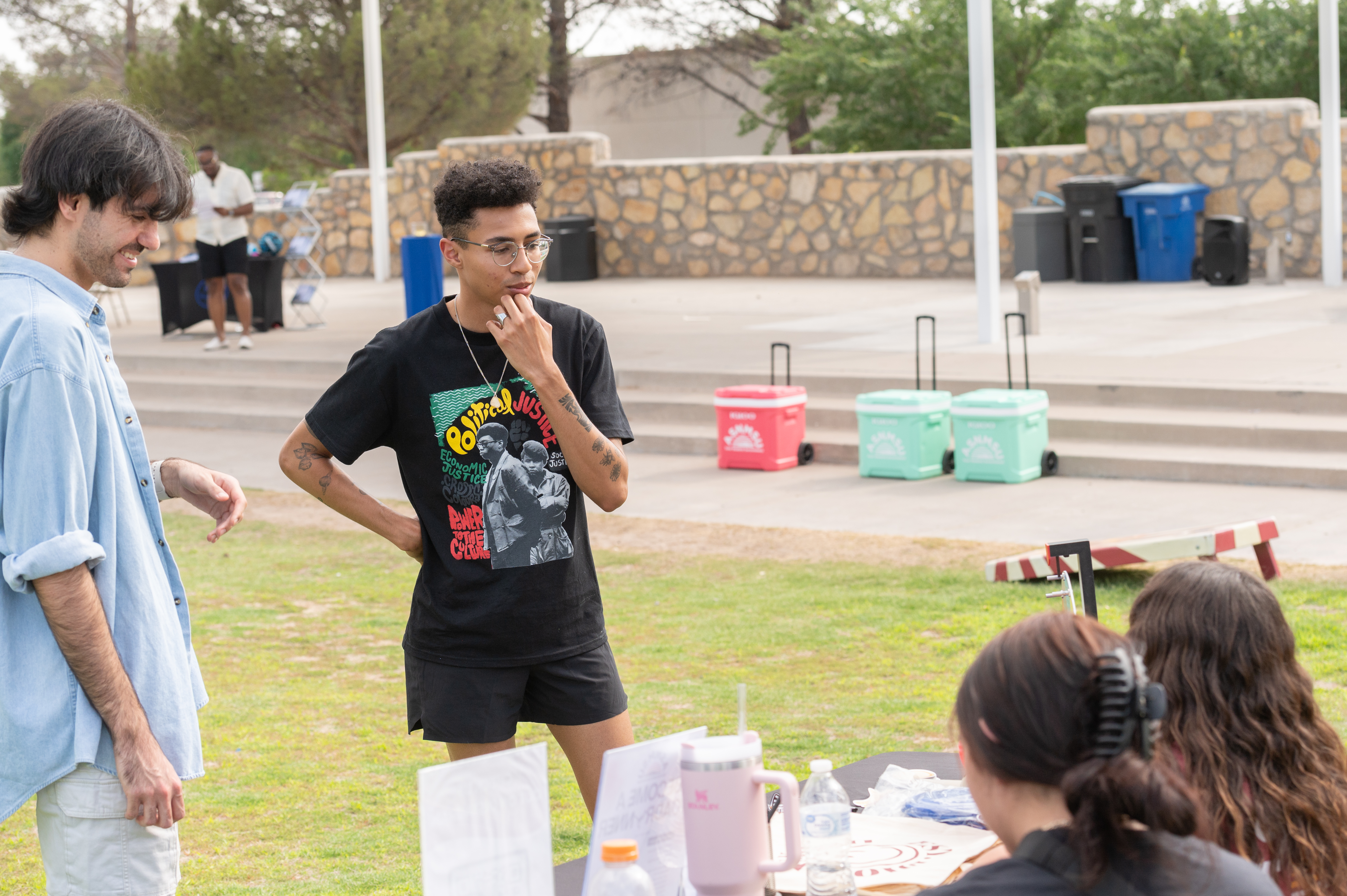 Picture from the celebration of Juneteenth, The image depicts an outdoor gathering with people interacting around a table set up on a grassy area. Two individuals, one in a light blue button-up shirt with rolled-up sleeves and the other in a black T-shirt and shorts printed with "Political Justice" and a graphic design, stand engaged in conversation in the foreground. They appear to be in a casual, friendly discussion. In the background, a stone wall is visible, along with a few blue, red, and turquoise wheeled coolers staged on a concrete platform beside some recycling bins and trash cans. In the lower right corner, the back of a third participant’s head, who has dark hair tied back and headphones around their neck, is seen. A fourth person with long, wavy hair is partially visible, facing the two standing individuals. The setting appears to be a campus or community event with a relaxed and open atmosphere.